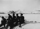 Sex flygsoldater sitter i snön och solar sig, cirka 1942. I bakgrunden syns fjället Kirunavaara, med gruvbyggnader.

Bildtext vid foto: 