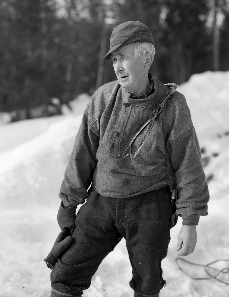 Tømmerkjører Kåre Bratteggen (1910-1999), fotografert under tømmerlunning på Lundsberget i Trysil vinteren 1977. Bildet er tatt etter avlessing av tømmeret på velteplassen. Hengende over skulderen har han tømmersaksa som han hadde brukt for å dra stokkene opp på tømmerlunna. Bratteggen var kledd i mørke vadmelsbukser og en grå busserull i halvull.