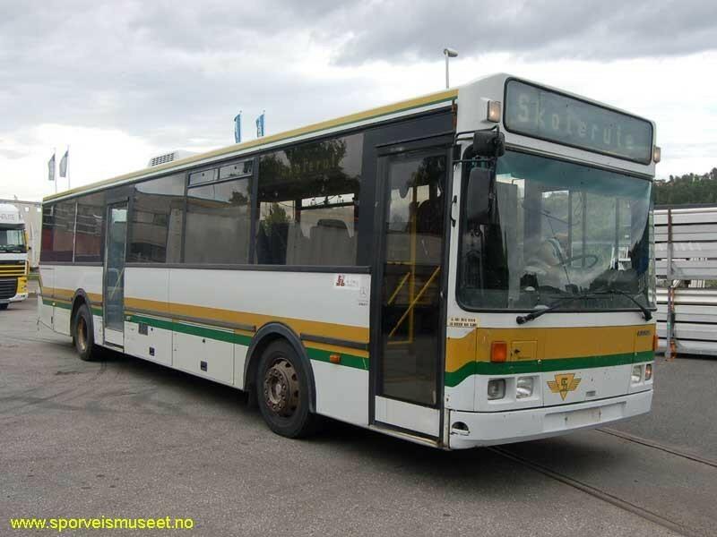Hvit buss med en gul og grønn stripe som går rundt bussen. Interiøret består av grått tak og gulv, og seter med et mønstret stoff i gått, blått og kremfarge. 