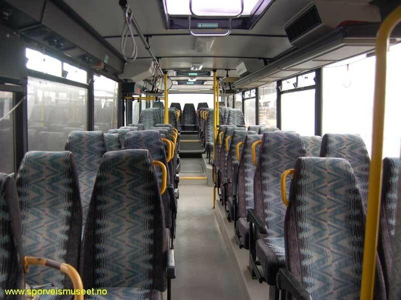 Hvit buss med en gul og grønn stripe som går rundt bussen. Interiøret består av grått tak og gulv, og seter med et mønstret stoff i gått, blått og kremfarge. 