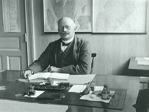 Troligen en järnvägstjänsteman som sitter vid sitt skrivbord. (Se kartan bakom med markerad järnvägssträckning.)