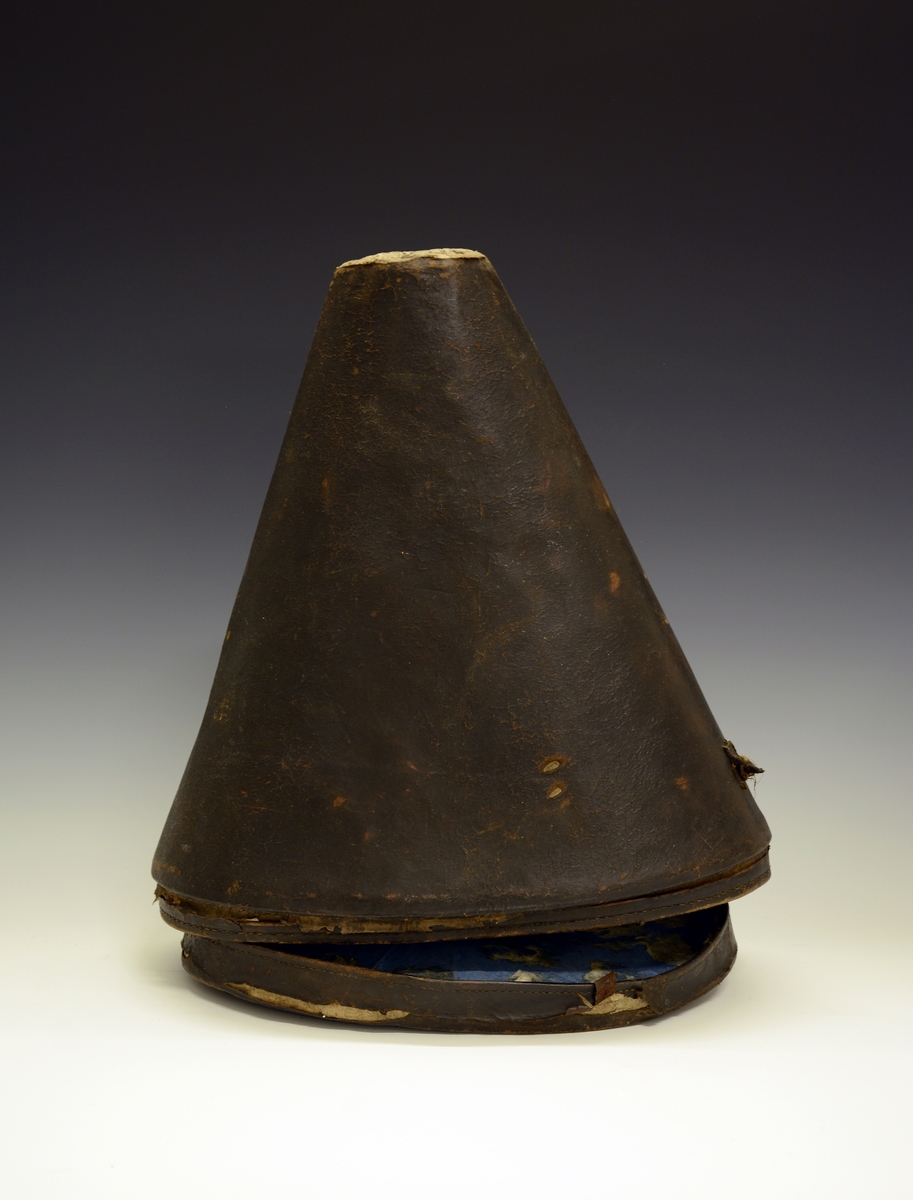 Hatteeske til pigghjelm. Pyramideformet med hull øverst. Utvendig er hatteesken av lær, innvendig er den av papp og polstret med vatt og fôret med hvitt linstoff med apperatur. Lokket er løst, og har et håndtak på hver side.