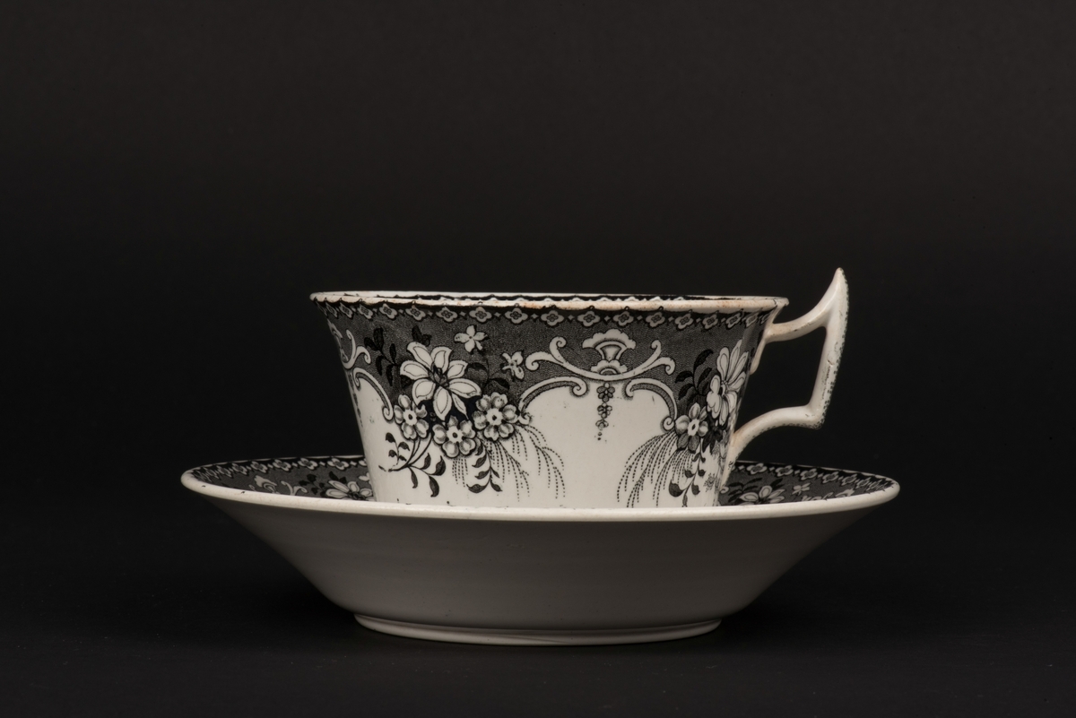 Kaffekopp med fat av vitt porslin med svart dekor.
Rik dekor på fatets ovansida och på koppens in- och utsida med blommor och dekorativa bårder. På fatens mitt och i botten på koppen ett par påfåglar.
Koppen vidgar sig mot mynningen och har ett rombiskt, lite utdraget öra.