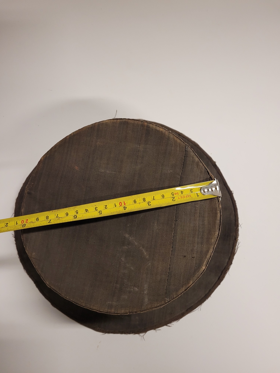 Hög hatt med brätten, överdragen med mörkt randigt linnetyg i fyra färger, övervägande brunt. Svettrem längs insidan av svart läder. Fodrad med vit lärft. Skullen uppbyggd av halm.