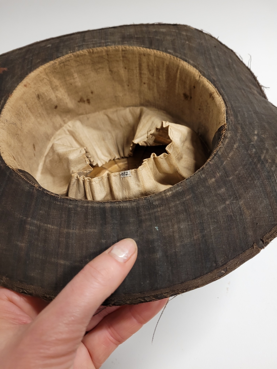 Hög hatt med brätten, överdragen med mörkt randigt linnetyg i fyra färger, övervägande brunt. Svettrem längs insidan av svart läder. Fodrad med vit lärft. Skullen uppbyggd av halm.