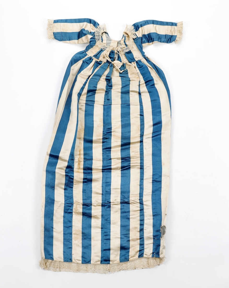 Dåpskjole i stoff med brede striper med blonder rund nedkant, ermer og halsringing. Håndsydd. Splitt bak , løpegang med snøring i nakken. Kjolen består av flere sammensydde stykker.