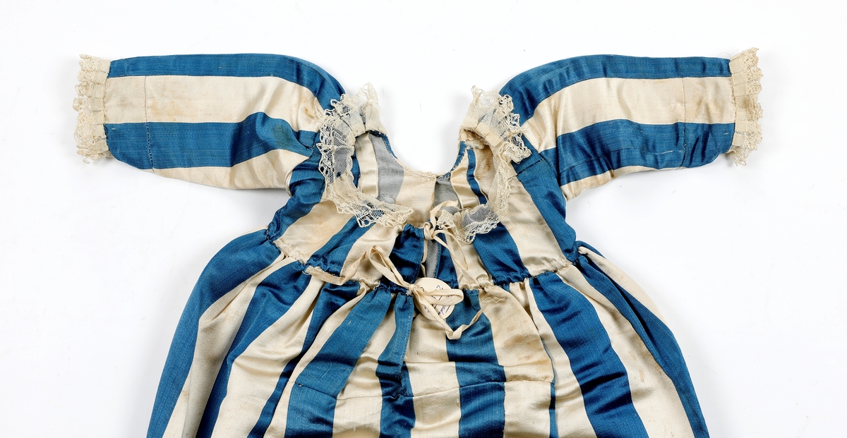 Dåpskjole i stoff med brede striper med blonder rund nedkant, ermer og halsringing. Håndsydd. Splitt bak , løpegang med snøring i nakken. Kjolen består av flere sammensydde stykker.