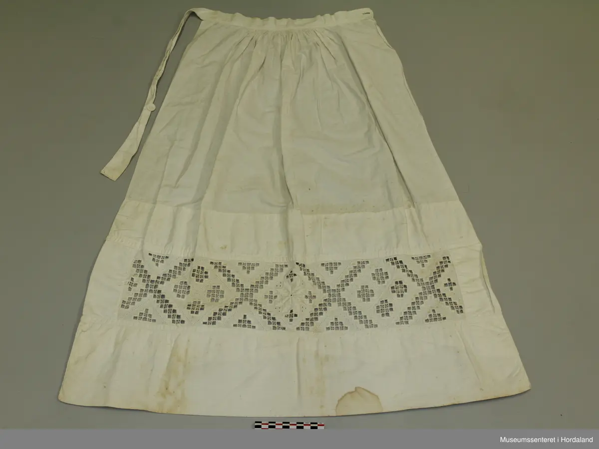 Kvitt bunadsforklede med hardangersaumbord felt inn 14 cm frå nedre kanten. Motivet er inndelt i firkantar, i midten framme er det sydd ei åttebladrose. Linning i livet som fortset rundt ryggen og knappast i sida. Forkledet er rynka ved linninga midt framme. Ikkje rynka i sidene.