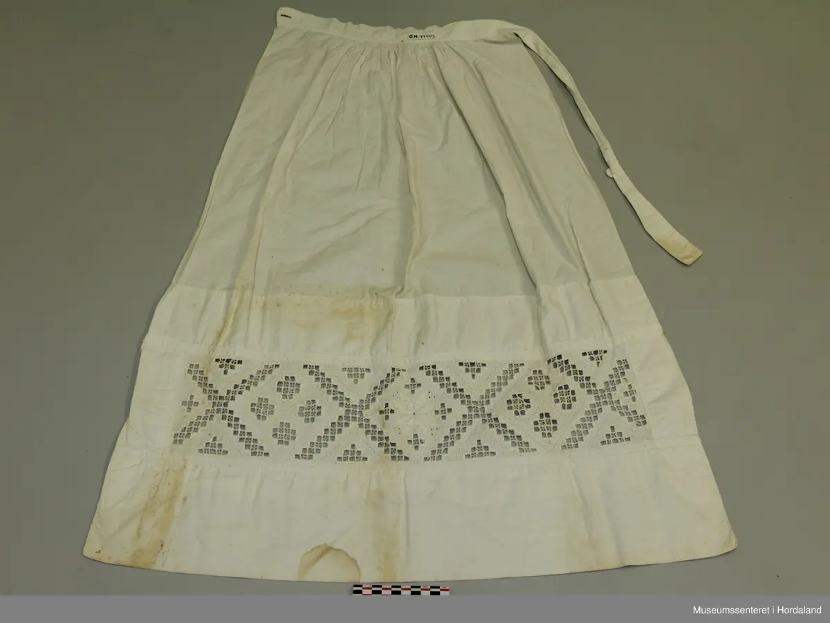 Kvitt bunadsforklede med hardangersaumbord felt inn 14 cm frå nedre kanten. Motivet er inndelt i firkantar, i midten framme er det sydd ei åttebladrose. Linning i livet som fortset rundt ryggen og knappast i sida. Forkledet er rynka ved linninga midt framme. Ikkje rynka i sidene.