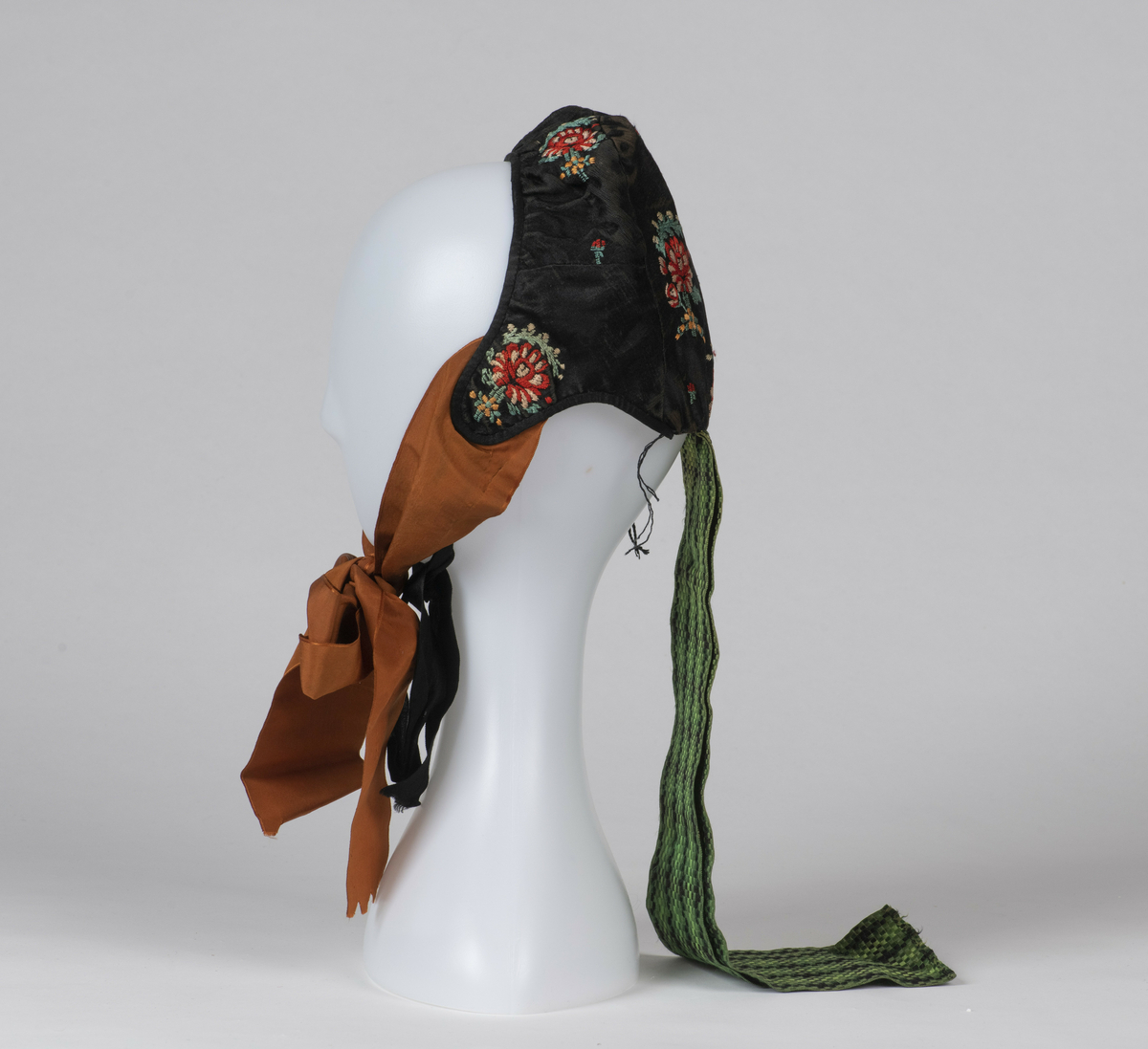 Lue. Kvinnelue, sydd av sort silke brodert med blomstermotiver. Brune knyttebånd av silke, samt nakkebånd vevd med ruter i sort og grønt. 