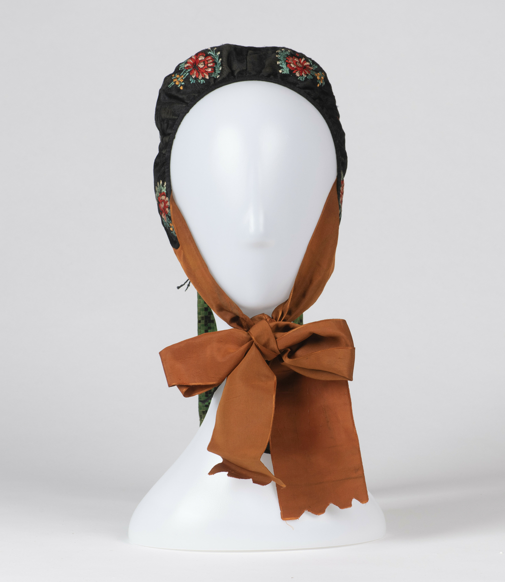 Lue. Kvinnelue, sydd av sort silke brodert med blomstermotiver. Brune knyttebånd av silke, samt nakkebånd vevd med ruter i sort og grønt. 