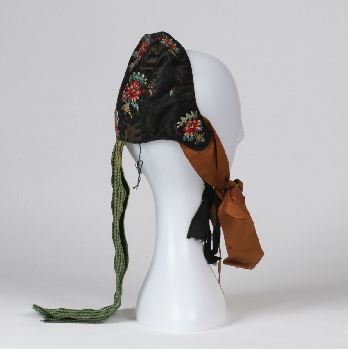 Lue. Kvinnelue, sydd av sort silke brodert med blomstermotiver. Brune knyttebånd av silke, samt nakkebånd vevd med ruter i sort og grønt. 