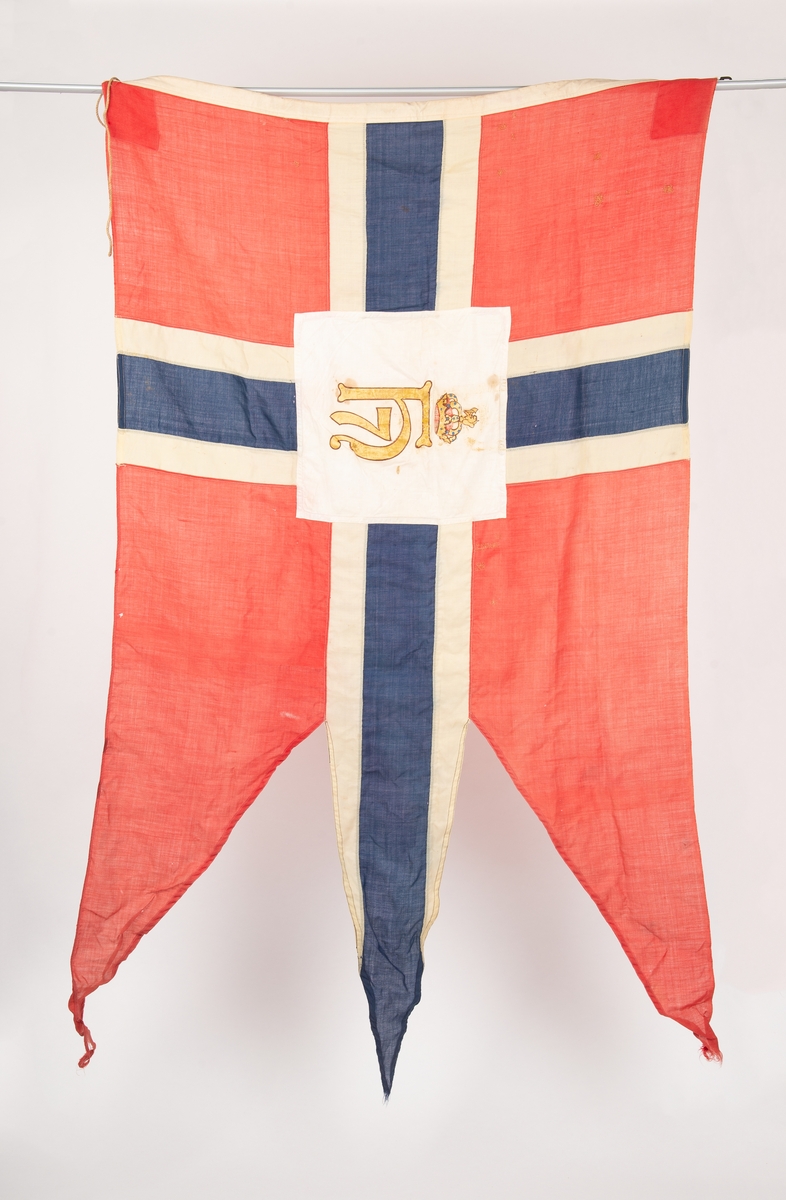 Norsk splittflagg med tunge. Hvit kvadratisk felt i midten med kong Haakon VII kronede monogram i gull og brunt. Tau i innerkanten med messingkrok. Diverse stoppede småhull.