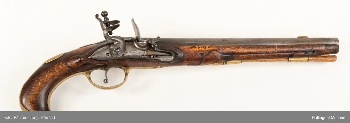 Flintlåspistol fra ca. 1750, fra tiden da Frederik V var konge over Danmark og Norge - se monogram på messingbeslag på skjeftet. Pistolskjeftet er utformet i tre med utskåret dekorasjon rundt låsen (begge sider og på oversiden) og under løpet. Har ladestokk som holdes fast av to messingbeslag. Kolbekappen og avtrekksbøylen er i messing. Mangler et messingbeslag foran under løpet.