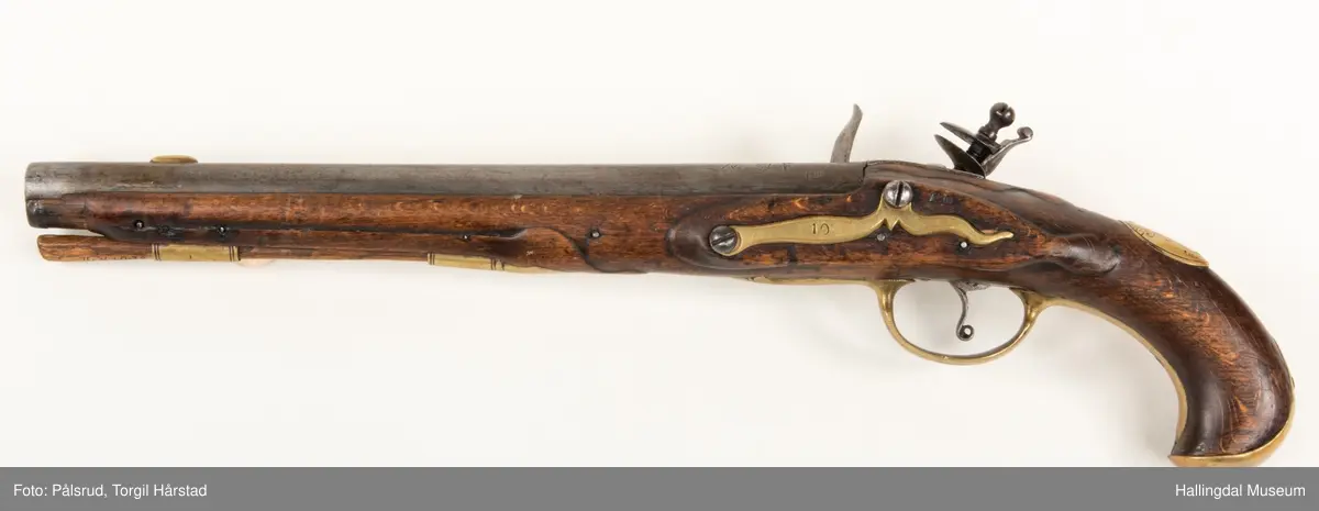 Flintlåspistol fra ca. 1750, fra tiden da Frederik V var konge over Danmark og Norge - se monogram på messingbeslag på skjeftet. Pistolskjeftet er utformet i tre med utskåret dekorasjon rundt låsen (begge sider og på oversiden) og under løpet. Har ladestokk som holdes fast av to messingbeslag. Kolbekappen og avtrekksbøylen er i messing. Mangler et messingbeslag foran under løpet.