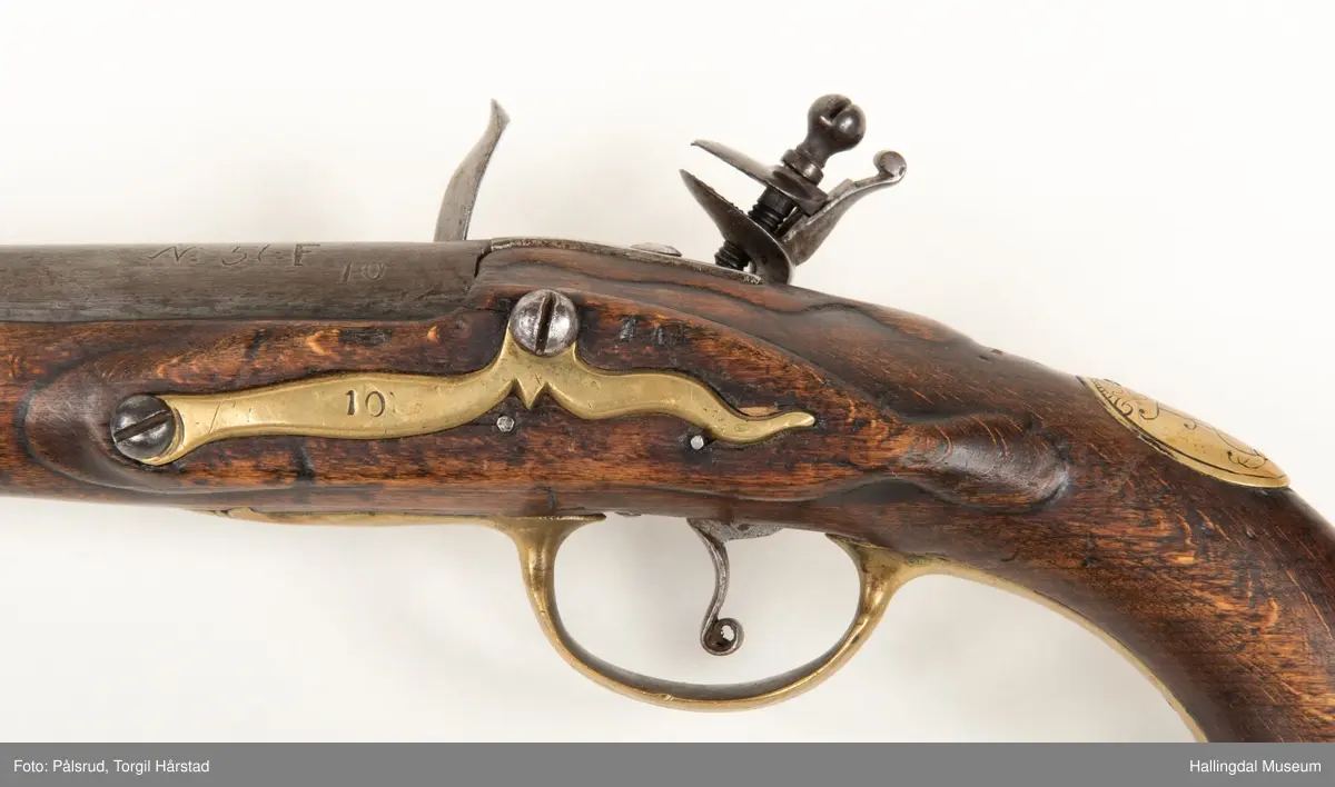 Flintlåspistol fra ca. 1750, fra tiden da Frederik V var konge over Danmark og Norge - se monogram på messingbeslag på skjeftet. Pistolskjeftet er utformet i tre med utskåret dekorasjon rundt låsen (begge sider og på oversiden) og under løpet. Har ladestokk som holdes fast av to messingbeslag. Kolbekappen og avtrekksbøylen er i messing. Mangler et messingbeslag foran under løpet.