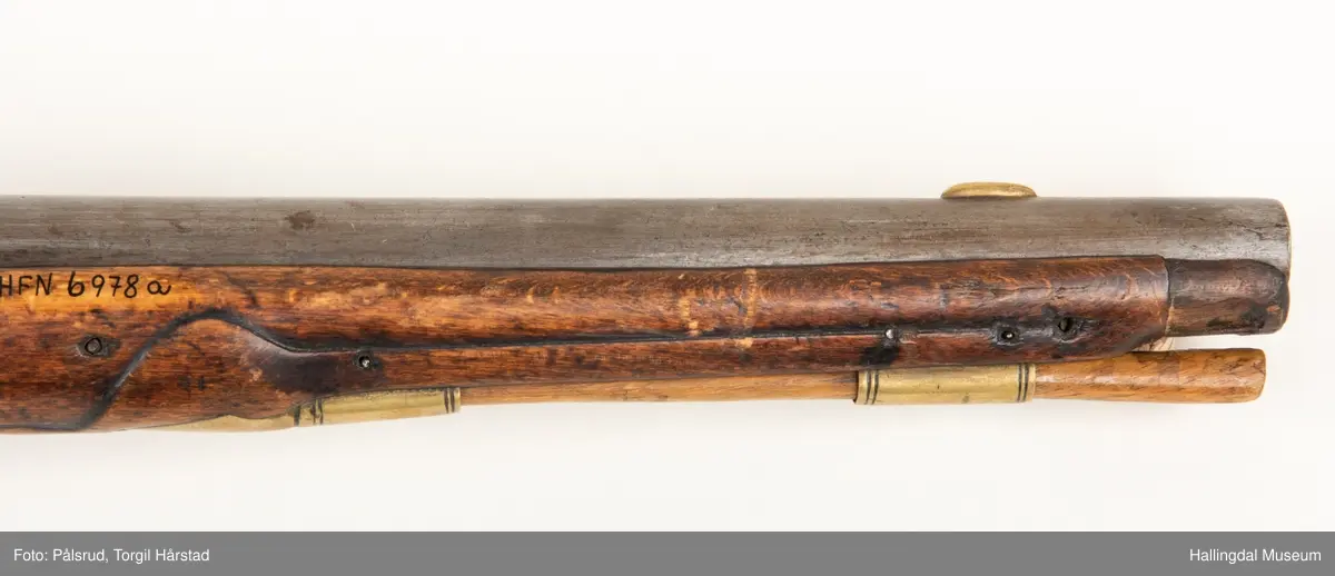 Flintlåspistol fra ca. 1750, fra tiden da Frederik V var konge over Danmark og Norge - se monogram på messingbeslag på skjeftet. Pistolskjeftet er utformet i tre med utskåret dekorasjon rundt låsen (begge sider og på oversiden) og under løpet. Har ladestokk som holdes fast av to messingbeslag. Kolbekappen og avtrekksbøylen er i messing. Mangler et messingbeslag foran under løpet.