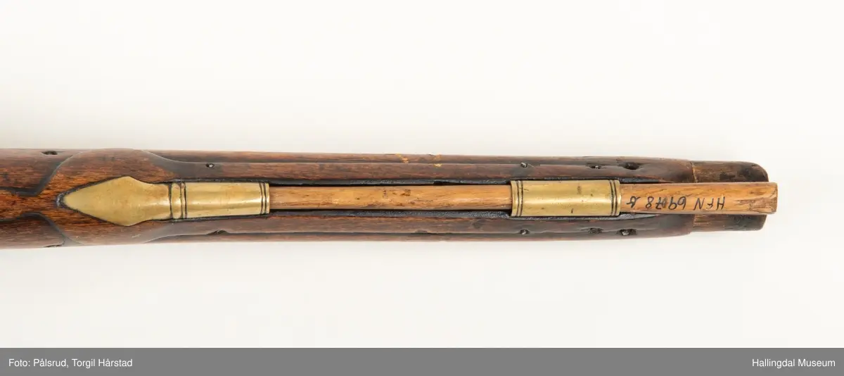 Flintlåspistol fra ca. 1750, fra tiden da Frederik V var konge over Danmark og Norge - se monogram på messingbeslag på skjeftet. Pistolskjeftet er utformet i tre med utskåret dekorasjon rundt låsen (begge sider og på oversiden) og under løpet. Har ladestokk som holdes fast av to messingbeslag. Kolbekappen og avtrekksbøylen er i messing. Mangler et messingbeslag foran under løpet.