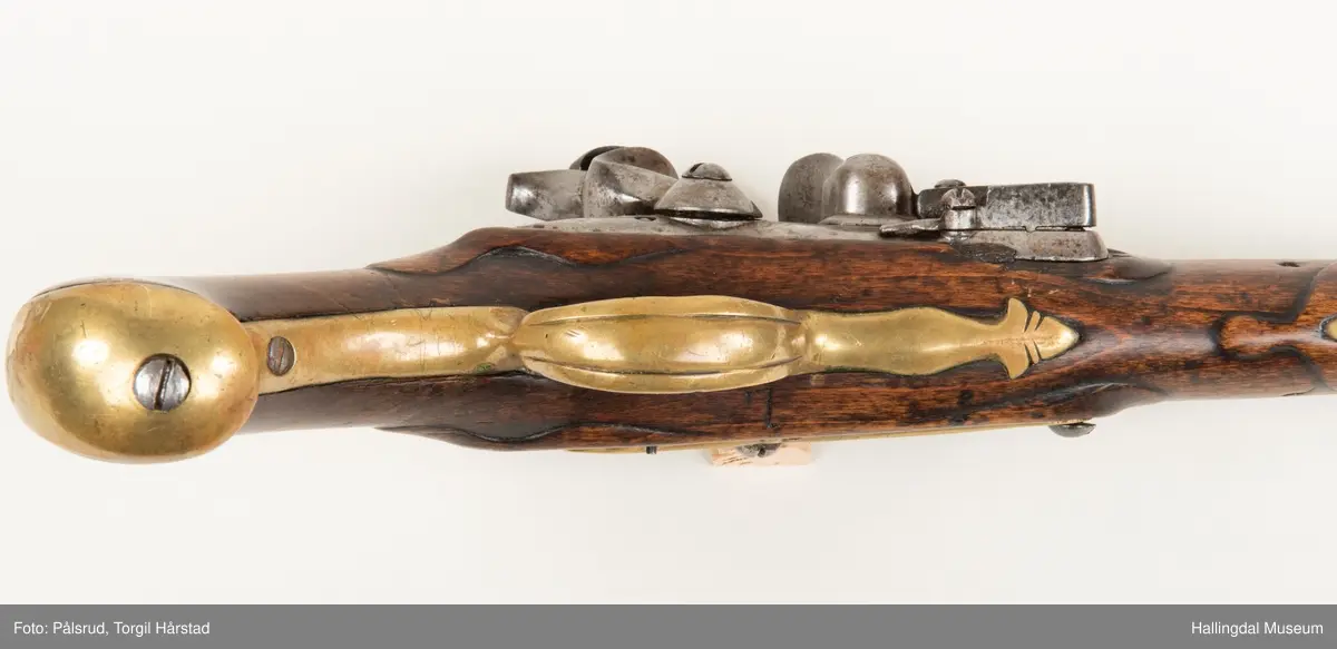 Flintlåspistol fra ca. 1750, fra tiden da Frederik V var konge over Danmark og Norge - se monogram på messingbeslag på skjeftet. Pistolskjeftet er utformet i tre med utskåret dekorasjon rundt låsen (begge sider og på oversiden) og under løpet. Har ladestokk som holdes fast av to messingbeslag. Kolbekappen og avtrekksbøylen er i messing. Mangler et messingbeslag foran under løpet.