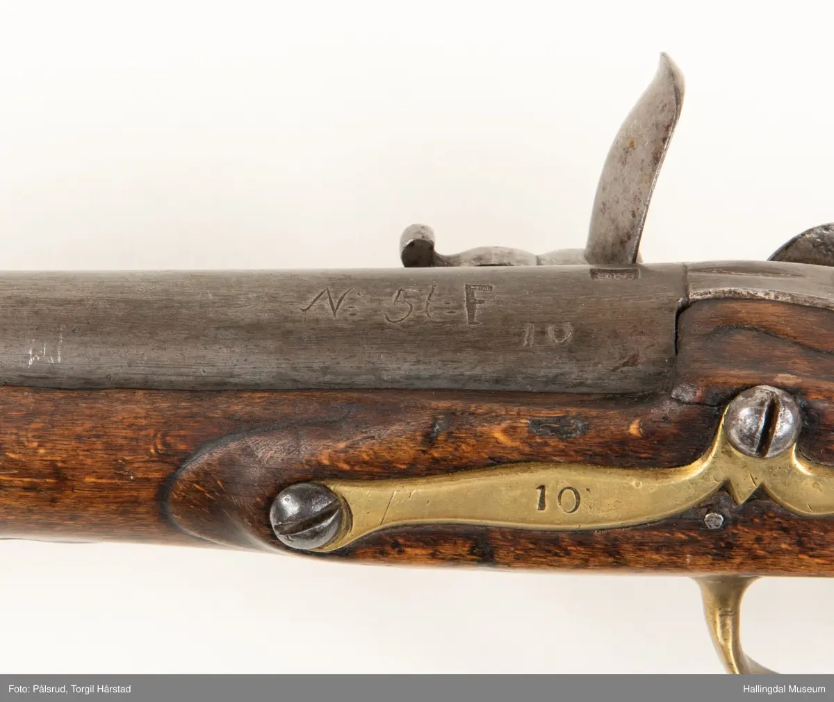 Flintlåspistol fra ca. 1750, fra tiden da Frederik V var konge over Danmark og Norge - se monogram på messingbeslag på skjeftet. Pistolskjeftet er utformet i tre med utskåret dekorasjon rundt låsen (begge sider og på oversiden) og under løpet. Har ladestokk som holdes fast av to messingbeslag. Kolbekappen og avtrekksbøylen er i messing. Mangler et messingbeslag foran under løpet.