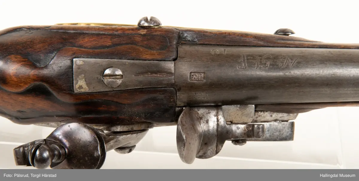 Flintlåspistol fra ca. 1750, fra tiden da Frederik V var konge over Danmark og Norge - se monogram på messingbeslag på skjeftet. Pistolskjeftet er utformet i tre med utskåret dekorasjon rundt låsen (begge sider og på oversiden) og under løpet. Har ladestokk som holdes fast av to messingbeslag. Kolbekappen og avtrekksbøylen er i messing. Mangler et messingbeslag foran under løpet.