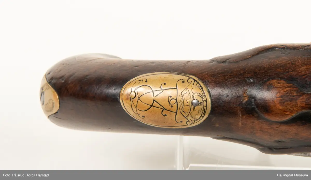 Flintlåspistol fra ca. 1750, fra tiden da Frederik V var konge over Danmark og Norge - se monogram på messingbeslag på skjeftet. Pistolskjeftet er utformet i tre med utskåret dekorasjon rundt låsen (begge sider og på oversiden) og under løpet. Har ladestokk som holdes fast av to messingbeslag. Kolbekappen og avtrekksbøylen er i messing. Mangler et messingbeslag foran under løpet.