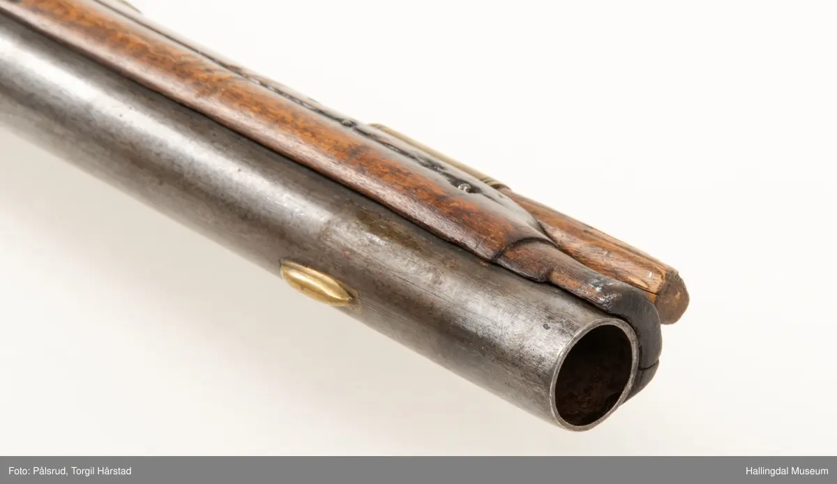 Flintlåspistol fra ca. 1750, fra tiden da Frederik V var konge over Danmark og Norge - se monogram på messingbeslag på skjeftet. Pistolskjeftet er utformet i tre med utskåret dekorasjon rundt låsen (begge sider og på oversiden) og under løpet. Har ladestokk som holdes fast av to messingbeslag. Kolbekappen og avtrekksbøylen er i messing. Mangler et messingbeslag foran under løpet.
