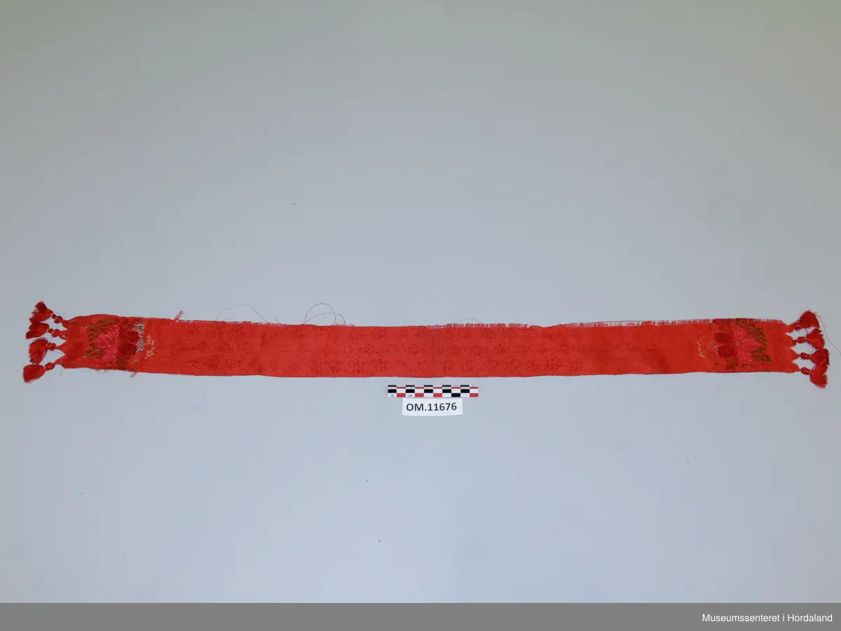 Smalt, lite silkeskjerf, til å knyta rundt halsen. Raud botnfarge med innvevd blomstermotiv. I kvar ende er det fire duskar og eit blomstermotiv med stor blomst og blad i fargane rosa, mørkeraud, grøn, lyseblå og gul. Litt flisete i kanten.