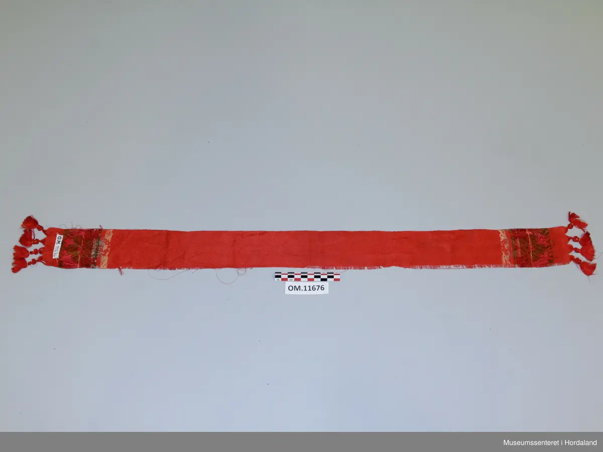 Smalt, lite silkeskjerf, til å knyta rundt halsen. Raud botnfarge med innvevd blomstermotiv. I kvar ende er det fire duskar og eit blomstermotiv med stor blomst og blad i fargane rosa, mørkeraud, grøn, lyseblå og gul. Litt flisete i kanten.