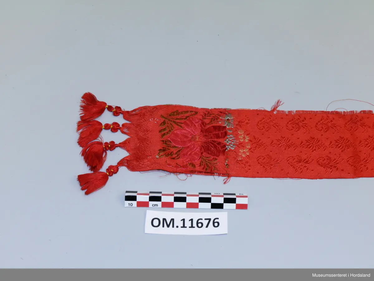 Smalt, lite silkeskjerf, til å knyta rundt halsen. Raud botnfarge med innvevd blomstermotiv. I kvar ende er det fire duskar og eit blomstermotiv med stor blomst og blad i fargane rosa, mørkeraud, grøn, lyseblå og gul. Litt flisete i kanten.