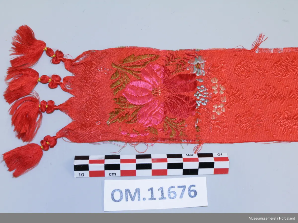 Smalt, lite silkeskjerf, til å knyta rundt halsen. Raud botnfarge med innvevd blomstermotiv. I kvar ende er det fire duskar og eit blomstermotiv med stor blomst og blad i fargane rosa, mørkeraud, grøn, lyseblå og gul. Litt flisete i kanten.