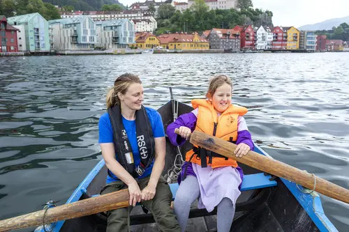 En voksen dame og en jente, begge med redningsvester, ror hver sin åre i en robåt på vannet ved Norges Fiskerimuseum