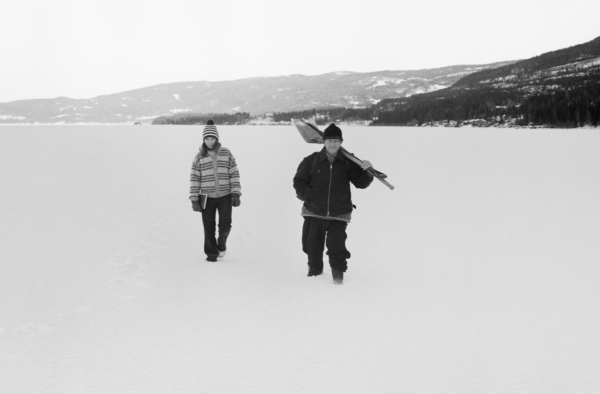 Gunhild Dæhlie og Paul Stensæter, fotografert på den snødekte isflata på Steinsfjorden, en sidearm til Tyrifjorden på Ringerike vinteren 1973. Stensæter bodde på østsida av Steinsfjorden, i Hole kommune, og han drev næringsfiske nesten hele året. Vinterstid var det garnfisket etter sik under isen som gav de største fangstene. Her var han på veg ut for å trekke garn, noe han gjorde med to døgns mellomrom. Han bar med seg ei isbile og en spade. Denne redskapen trengte han, for garna sto mellom hull i isen som oftest fikk ei isskorpe og noen ganger også var gjendrevet med snø, mellom hver gang garna skulle etterses. Gunhild Dæhlie var gift med fotografen som tok dette bildet, museumsmannen Åsmund Eknæs. Mens Åsmund tok bilder bar Gunhild feltdagboka hans. 

I 1970-åra var etnologen Åsmund Eknæs fra Norsk Skogbruksmuseum flere ganger på besøk hos Paul Stensæter for å observere ham i aktivitet som fisker og intervjue ham om denne virksomheten. Eknæs oppsummerte det han fikk se og høre om garnfisket under isen på Steinsfjorden slik:

«Sikfiske med garn. Dette fisket foregikk på to steder, i åpent vann ute i Tyrifjorden og under isen i Steinsfjorden. Tyrifjorden er ofte åpen langt utover vinteren og det hender at den ikke legger seg i det hele tatt. De dro da i båt over fra Steinsfjorden og satte garn på ganske store dyp, 20-40 favner. Når det ble fisket for fullt ble det brukt 20 garn. 10 sto ute mens de øvrige var hjemme til tørking.

Garnfisket under isen i Steinsfjorden begynner så fort isen legger seg om høsten, fordi tynn og gjennomsiktig is er en fordel når garna skal settes ut første gang. Å sette ut garn under isen kaller Paul for øvrig «å høgge ut garna». Dette krever en spesiell teknikk. Er isen gjennomsiktig, foregår det på følgende måte: To hull hogges med så lang avstand som lengda på garnet. Ei rett granstang på 7-8 m stikkes ned i det ene hullet med den tynneste enden først. I den tykkeste enden er det et hull hvor det blir festet ei snor. Stanga blir nå skjøvet i full fart mot det andre hullet. En viktig detalj ved denne stanga er at den skal være nyhogget. Da ligger den dypere i vannet og skubber mindre mot isen. Stanga går ikke helt fram til det andre hullet. Der den stopper blir det hogget et mindre hull og den skyves videre ved hjelp av en kjepp med ei kløft i enden. Når snora er brakt fram på denne måten, er det en enkel sak å trekke garnet under isen. 

Men er isen ugjennomsiktig, slik at det er umulig å se stanga, må Paul gjøre det på en annen måte. Da finner han ei lang stang med god krumming på. Så hogger han hull så tett at stanga kan stikkes ned i det ene og komme opp igjen gjennom det neste.

Garnet er nå på plass under isen og står på bunnen på 5-10 favners dyp. Fra hver ende av garnet går det ei tynn snor opp til hullet i isen. Snorene går ikke opp gjennom hullet, men er festet litt ved siden. Dette er gjort for å hindre at han hogger dem av når is som har dannet seg i hullet skal fjernes.

Dagens nylongarn tåler å stå ute hele vinteren. Tidligere, da lin og bomull var mest brukt, var det nødvendig å ta garna opp og tørke dem. Av de 20-40 garn som Paul brukte, var halvparten til tørk.

Ettersynet, som gjerne foregår annenhver dag, begynner med at han får tak i snorene fra garnet ved å stikke en pinne med krok på innunder isen. I den borteste enden løsner han garnsnora og fester isteden ei lang nylonsnor til garnet. Tidligere brukte han snor av tvunnet hestetagl. Snora er så lang at den også rekker bort til der han står oppå isen. Ved trekkinga tar han ut fisken etter hvert. Når hele garnet er trukket, blir han stående på samme sted, tar tak i snora og drar garnet ut igjen samtidig som han passer på at det går ordentlig.

I sterk kulde vil det våte garnet fryse til en klump og være umulig å sette igjen. For å hindre dette hogger han ei grop i isen bak hullet og fyller denne med vann. Etter hvert som han trekker garnet putter ha det ned i gropa og det holder seg opptint til det skal settes igjen.

Når det blir mildvær og fare for at isen skal gå opp og komme i drift, må det tas spesielle forholdsregler for å hindre at garna blir dratt med isflak og forsvinner. Paul binder i slike tilfelle inn en bit snelletråd i den snora som går fra isen og ned til garnet. Kommer isen i drift, vil snelletråden ryke, og garnet blir stående igjen på samme sted. Det er da en forholdsvis enkel sak å sokne etter det.

Garnfisket ga jevne tilførsler av sik gjennom det meste av vinteren. Vintersiken i Steinsfjorden er noe mindre enn høstsiken. Det går omkring 4 på kiloen, mot 3 om høsten. Fangstene varierer fra noen få til 25-30 pr. garn. Med 10 garn ute vil han kunne få opptil 200-300 sik eller 50-70 kilo. Men da blir garnene trukket bare annenhver dag.»