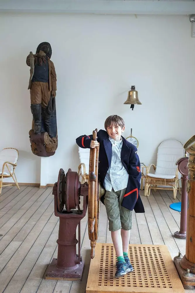 En gutt ved Skipsrattet. Det er en bjelle og en figur med noen eldre klær i bakgrunnen