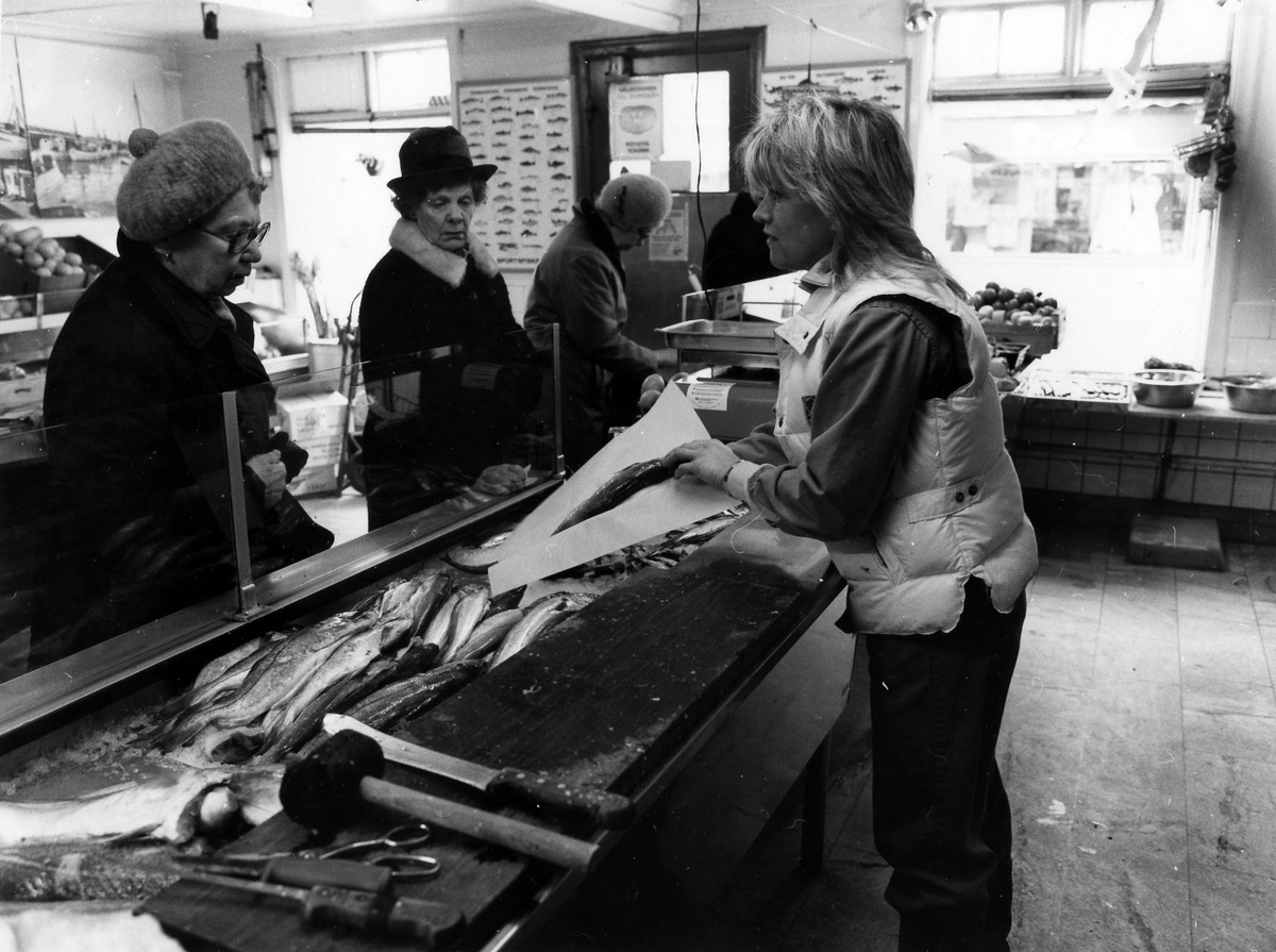 Kvinna bakom disken betjänar kunder på Västkustens fiskaffär. Fastigheten revs i månadsskifter juli-augusti 1984.