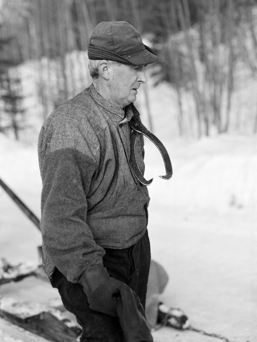 Tømmerkjører Kåre Bratteggen (1910-1999), fotografert under tømmerlunning på Lundberget i Trysil vinteren 1977. Bildet er tatt etter avlessing av tømmeret på velteplassen. Hengende over skulderen har han tømmersaksa som han hadde brukt for å dra stokkene opp på tømmerlunna. Bratteggen var kledd i mørke vadmelsbukser og en grå busserull i halvull.