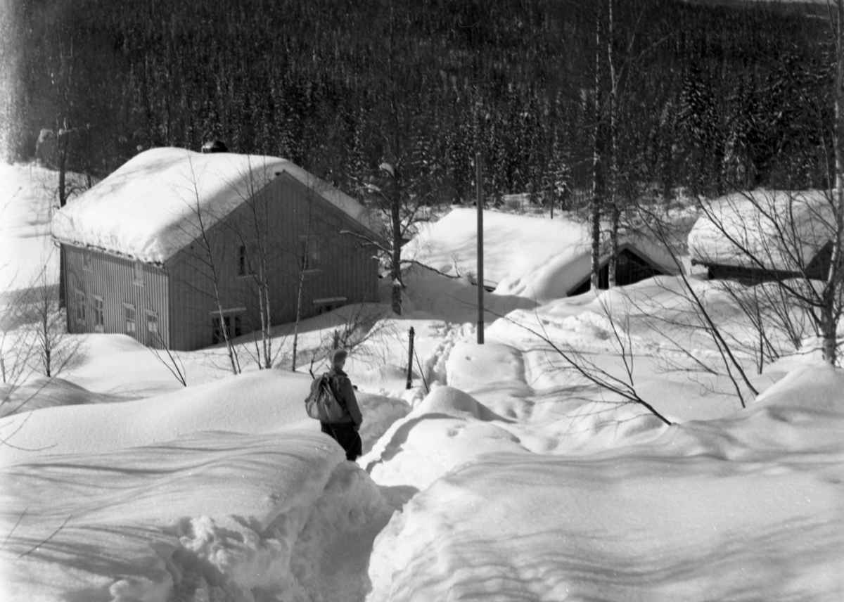 Vinter med mye snø. En opptråkket vei hvor en mann står og titter mot et hus og flere bygninger.