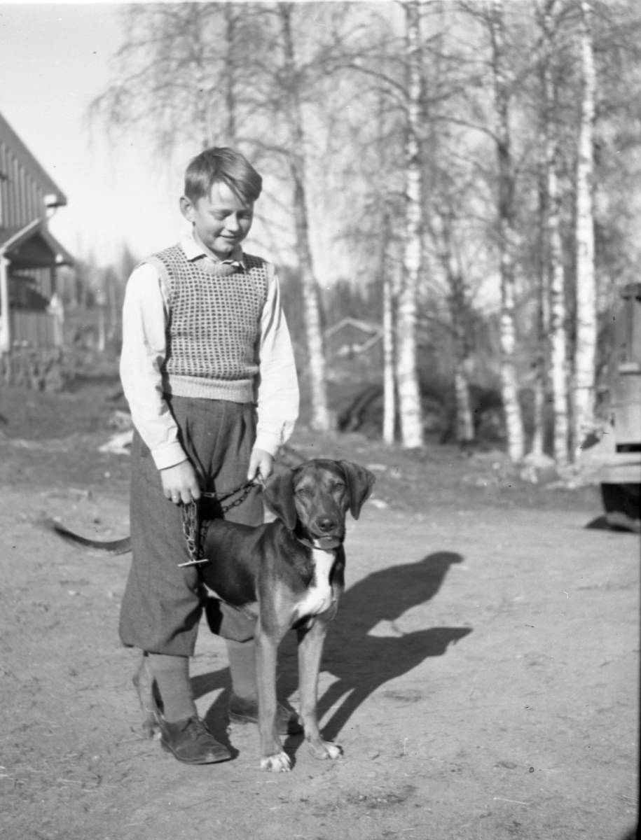 En gutt med en hund stående på gårdsplass. Hus kan skimtes i bakgrunnen.