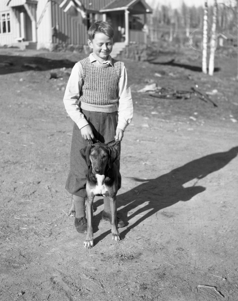 En gutt med en hund stående på gårdsplass. Et hus kan skimtes i bakgrunnen.