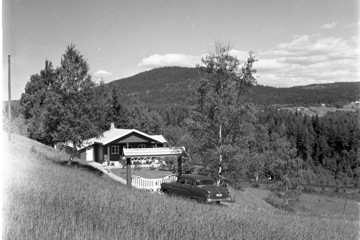 Landskapsbilde. En ås med bygninger sees i bakgrunnen. Et hus litt til venstre i bildet. Foran inngangsporten til huset står det parkert en bil.