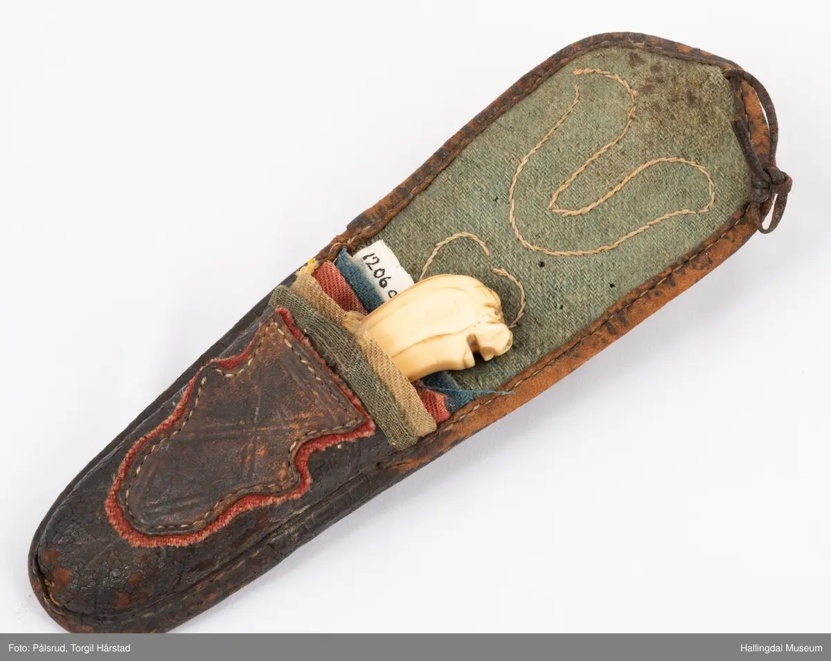 Kniv med etui. 
Etuiet er laget av mørkebrunt lær og ullstoff i grønt, rødt, gult og blått. Den er en tilnærmet rektangulær form som er avrundet i bunn og lokk. Er sydd sammen av to stykker med en ytterside og en innerside. Yttersiden er av mørkebrunt lær med påsydd lærkant. Den bredeste delen kan brettes over som lokk og har en utskjært hjerte- og en utskjært tulipanform. Lokket har på innsiden en lærreim som feste. Begge formene er kantet med lyse sting og foret med rødt ullstoff. 
På innsiden er etuiet foret med grønt ullstoff der de lyse stingene fra formene på lokket er synlig. kniven oppbevares i en av fire lommer som er kantet med grønt, gult, rødt og blått ullstoff. Selve yttersiden av etuiet/eller knivsliren er av mørkebrunt lær, kantet med samme lærkant som yttersiden og lokk, og er dekorert med linjer på sidene. Den er også dekorert med en påsydd lærlomme som er foret med rødt ullstoff som synes på kantene. Sydd med lyse sting og er preget med med et kryss og en V. 

Kniven har et skaft med et utskåret dyrehode (hvalross) og et laget av en hvalrosstann. Noe skadd. Knivbladet er festet gjennom hele lengden av skaftet, er rustent og har avbrekte deler på knivbladet. Knivblad stemplet med P. C./eller O. Westlander.