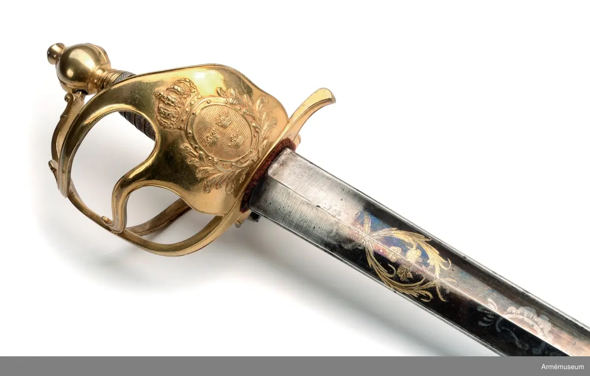 Fäste av förgylld mässing med två yttre och en inre sidobygel. Parerplåten har en krönt sköld omgiven av stiliserade palmkvistar. Kaveln klädd med hajskinn och lindad med en tvinnad,  förgylld mässingtråd. 

Klingan är rak och tveeggad. Bredd vid fästet: 37 mm. Upptill rester av blånering och dekorerad med tre kronor, omslutna av två palmkvistar.