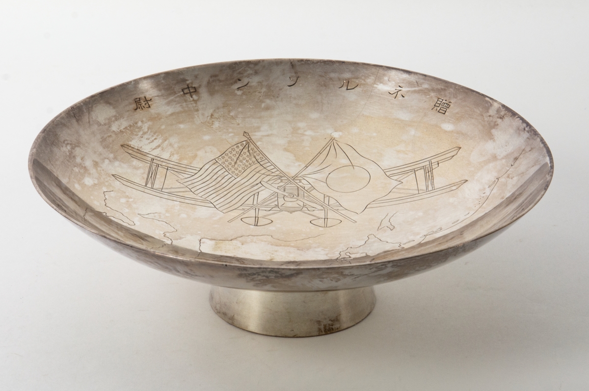 Fat på fot. Fatet har ett ingraverat motiv av flygplanet Douglas World Cruiser med två korsade flaggor framför, japanska och amerikanska. Under planet en karta som visar flygrutten för "Around the World Flight" 1924. Runt fatets insida är japanska skrivtecken ingraverade.