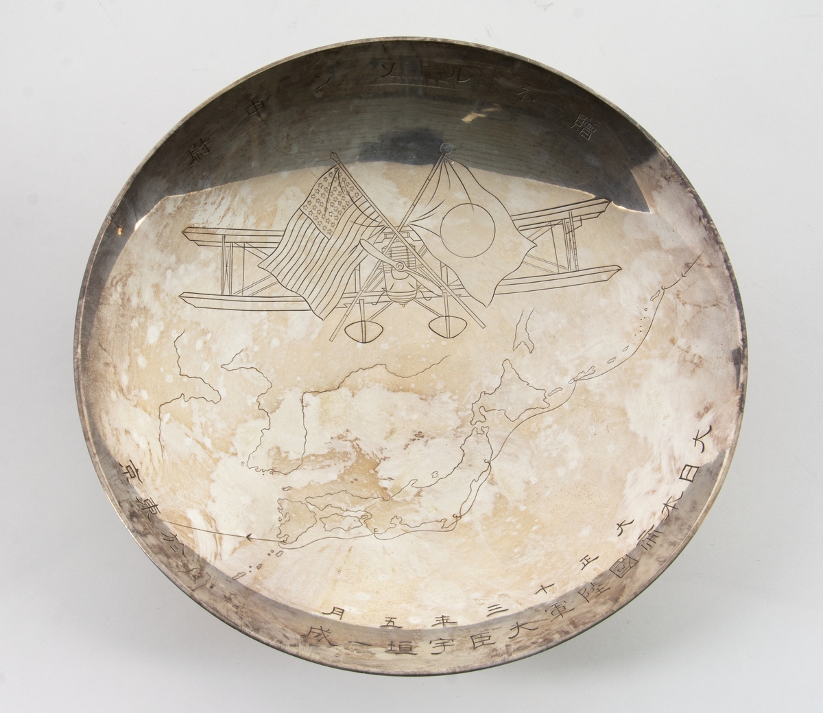 Fat på fot. Fatet har ett ingraverat motiv av flygplanet Douglas World Cruiser med två korsade flaggor framför, japanska och amerikanska. Under planet en karta som visar flygrutten för "Around the World Flight" 1924. Runt fatets insida är japanska skrivtecken ingraverade.