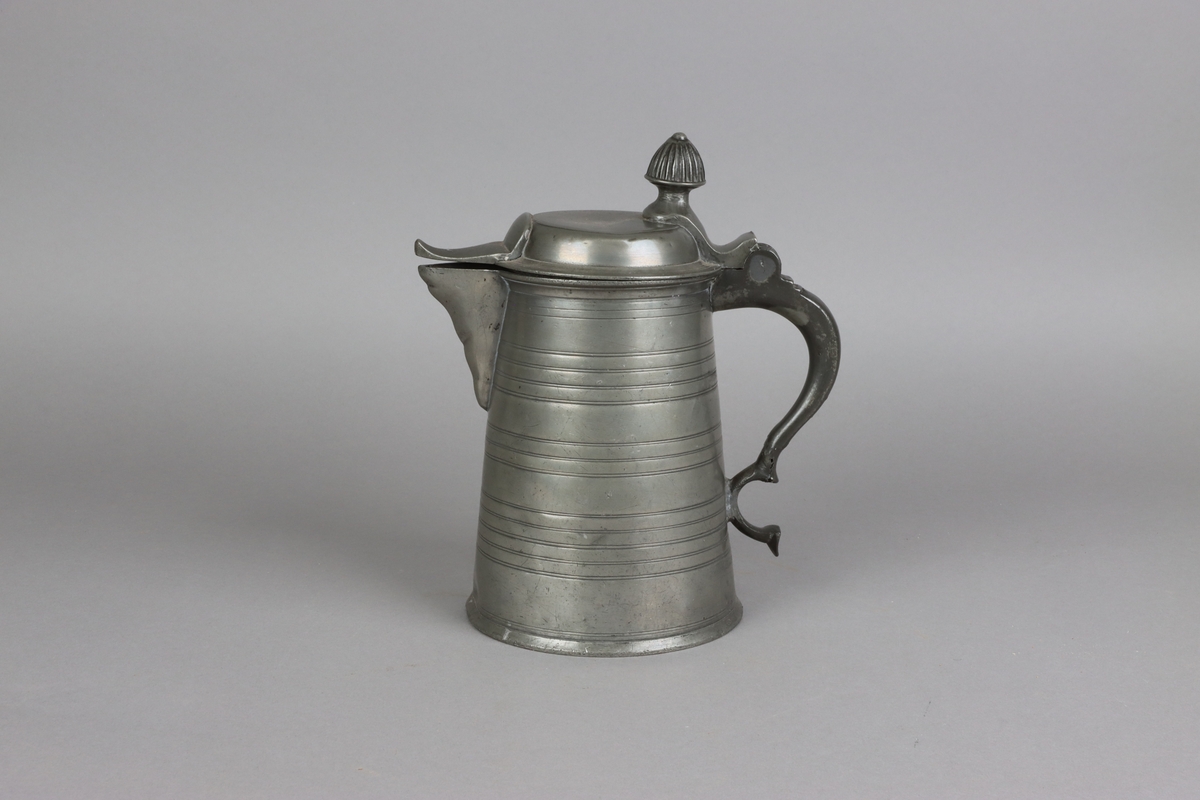 Jug and cover, all in pewter, with conical body  and a loop handle. On the cover knob in the form of a pine cone. On the body sections of horizontal borders. Five hallmarks inside the jug.

