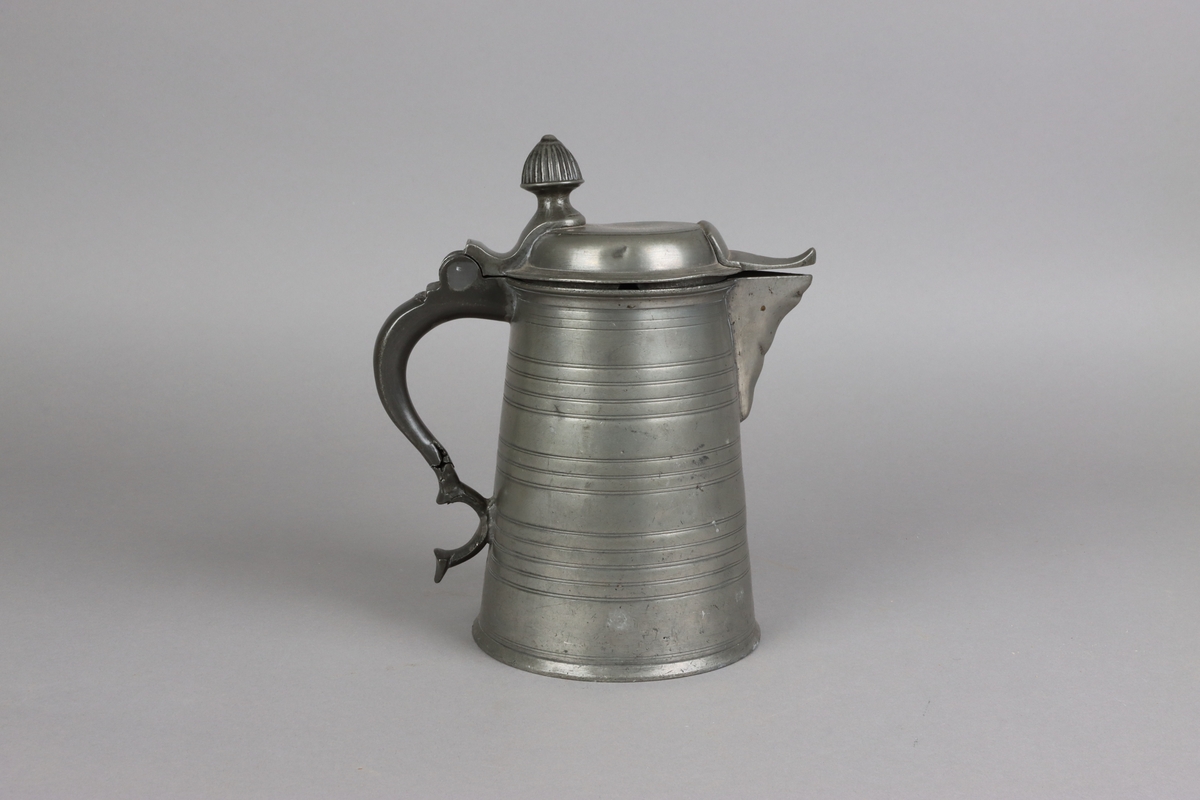 Jug and cover, all in pewter, with conical body  and a loop handle. On the cover knob in the form of a pine cone. On the body sections of horizontal borders. Five hallmarks inside the jug.
