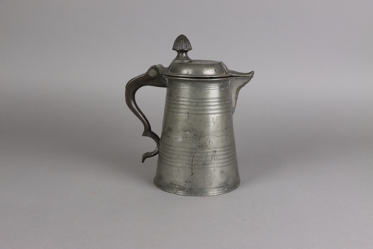 Jug and cover, all in pewter, with conical body  and a loop handle. On the cover knob in the form of a pine cone. On the body sections of horizontal borders. Five hallmarks inside the jug.