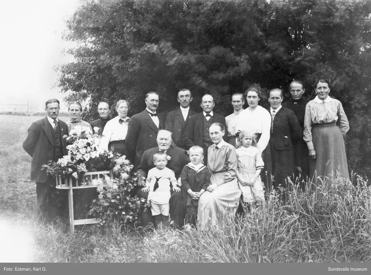 Gruppfoto utomhus, troligen någon bemärkelsedag. Sittande framför de andra är garvare Per Gustav Mattias Eckman och hans hustru Kristina Regina i Häre, Indal. Övriga vuxna och barn är oidentifierade. Ur Karl Eckmans samling.