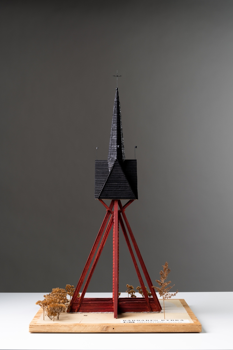 Modell av klockstapel på Barnarps kyrkogård, av trä på kvadratisk bottenplatta med plexiglas som sidor och tak. Runt klockstapeln finns stiliserad buskvegetation som utgörs av bland annat torkad rölleka (åga växter). Klockstapeln har tre hjärtstolpar och åtta stödben med dubbla strävor placerade på syllar. Allt är rödfärgat. Klockstapeln har ett svartfärgat korstak med åttakantig spira. På gavelspetarna sitter vindflöjlar och på spiran ett kors med fyra armar. Tre klockor hänger under korstaket. På bottenplattan sitter en etikett med text:"BARNARPS KYRKA FÖRSLAG TILL RESTAURERING AV KLOCKSTAPELN MODELL / SKALA 1:50 1973-07-28 L+M ARKITEKTERNA LÖFSTEDT+MOESCHLIN AB".