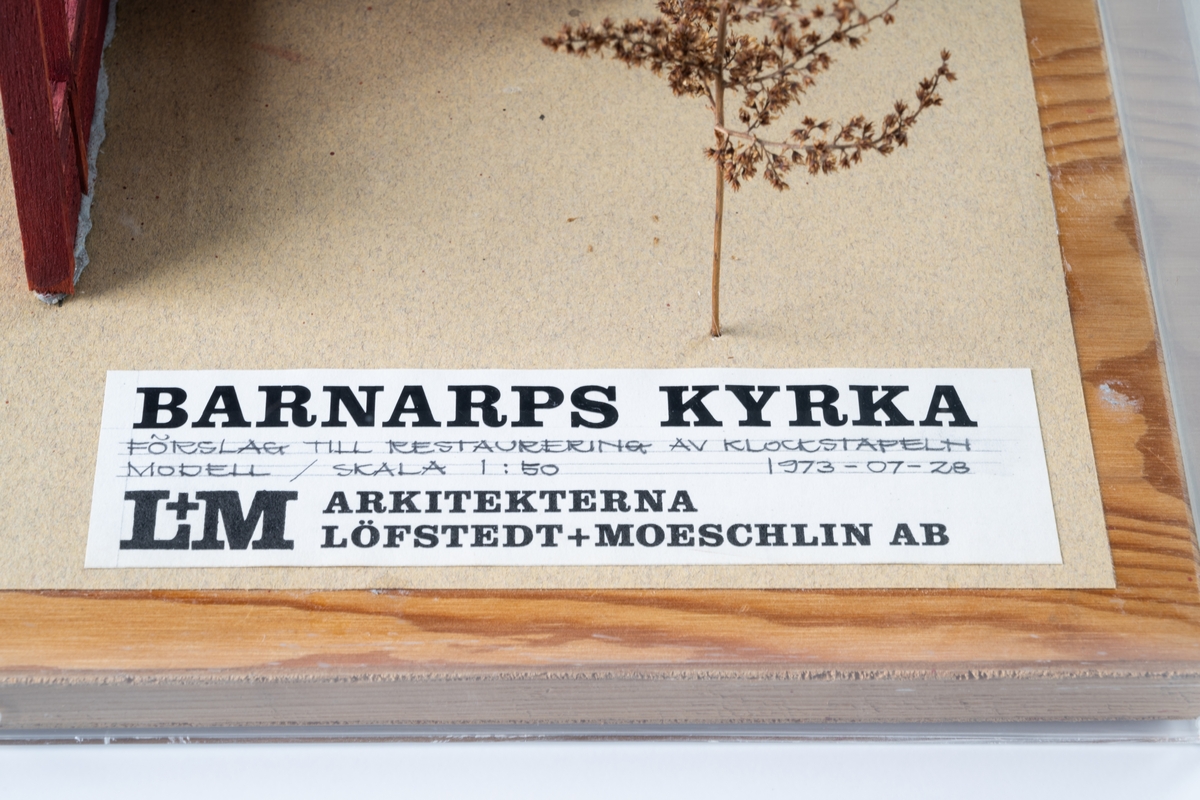 Modell av klockstapel på Barnarps kyrkogård, av trä på kvadratisk bottenplatta med plexiglas som sidor och tak. Runt klockstapeln finns stiliserad buskvegetation som utgörs av bland annat torkad rölleka (åga växter). Klockstapeln har tre hjärtstolpar och åtta stödben med dubbla strävor placerade på syllar. Allt är rödfärgat. Klockstapeln har ett svartfärgat korstak med åttakantig spira. På gavelspetarna sitter vindflöjlar och på spiran ett kors med fyra armar. Tre klockor hänger under korstaket. På bottenplattan sitter en etikett med text:"BARNARPS KYRKA FÖRSLAG TILL RESTAURERING AV KLOCKSTAPELN MODELL / SKALA 1:50 1973-07-28 L+M ARKITEKTERNA LÖFSTEDT+MOESCHLIN AB".